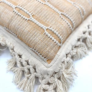 May include: A square throw pillow featuring a textured tan and beige woven fabric with vertical white stitched lines. The edges of the pillow are finished with a thick, cream-colored border adorned with decorative knotted fringe tassels.