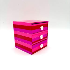 May include: A small pink and red striped dresser with three drawers. Each drawer has a white flower-shaped knob.