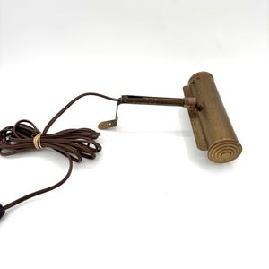 May include: A brass picture light with a long, adjustable arm and a brown cord. The light has a cylindrical shade with a ribbed design.