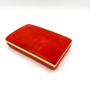 May include: A rectangular, red velvet box with gold edging. The box is closed and has a smooth, plush texture. The gold edging adds a touch of elegance. Suitable for jewellery or small keepsakes. The box is approximately 15 cm long.