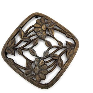 May include: A square-shaped metal trivet with rounded corners, featuring an openwork design of two daisy-like flowers connected by stems and leaves. The metal has a dark, aged bronze finish with visible patina and texture throughout the surface.