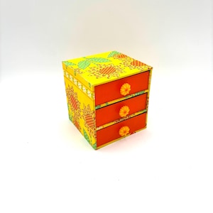 May include: A small, three-drawer jewelry box. The box is yellow with a floral pattern and orange drawers. Each drawer has a yellow flower-shaped knob. The box is a decorative storage item.