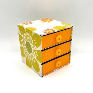 May include: A small, three-drawer dresser with a floral print on the exterior. The drawers are orange and have yellow flower-shaped knobs.