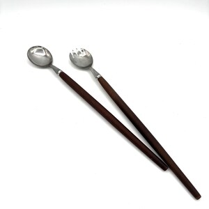 May include: Two serving spoons with stainless steel heads and dark brown wooden handles. One spoon is solid, the other has tines. The spoons are angled against a white background. The spoons are likely for serving salad or other dishes.