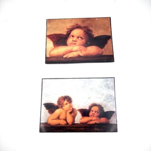 May include: Two rectangular wall plaques featuring classical art prints of cherubs. The top plaque shows a single winged cherub resting its chin on its hands. The bottom plaque displays two winged cherubs with one resting its chin on its hand.