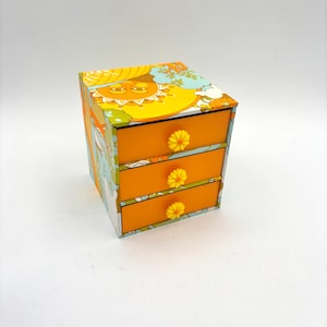 May include: A small, three-drawer chest of drawers with a retro, psychedelic design. The drawers are orange and have yellow flower-shaped knobs. The chest is covered in a colourful pattern of suns, flowers, and swirls.