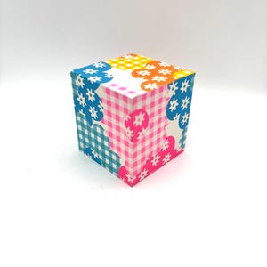 May include: A small, colourful cube with a patchwork design. Each side of the cube features a different pattern, including gingham, polka dots, and floral prints. The cube is made of cardboard and is approximately 7.5 cm in size.