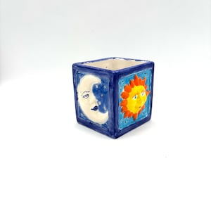 May include: A square ceramic planter featuring hand-painted celestial motifs. One side displays a white crescent moon against a dark blue background, while the adjacent side shows a bright yellow sun with orange rays on a light blue sky.