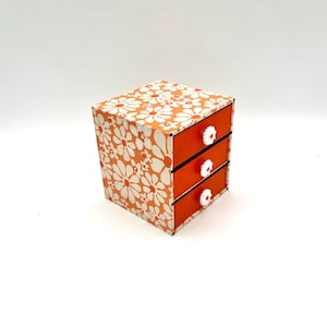 May include: A small orange and white floral patterned wooden box with three drawers. Each drawer has a white knob.