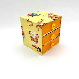 May include: A small, three-drawer wooden chest with a yellow background and orange drawers. The chest is decorated with a repeating pattern of cartoon tigers wearing flower crowns. The drawer pulls are yellow flower-shaped knobs.