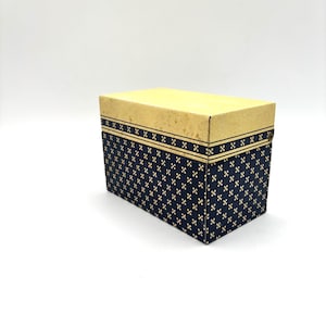 May include: A rectangular box with a light tan lid and navy blue sides. The sides feature a repeating pattern of small, gold-colored crosses. A gold band with a repeating pattern of small crosses and lines borders the top of the navy blue sides.