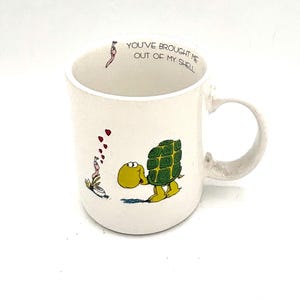 May include: White ceramic mug with a cartoon turtle design. The turtle is yellow and green, with a green shell. The mug has the text "YOU'VE BROUGHT ME OUT OF MY SHELL" printed on the inside rim.