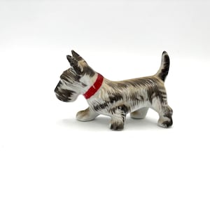 May include: A ceramic figurine of a Scottish Terrier dog. The dog has a black and white coat, a red collar, and is posed in a walking stance. The figurine is on a white background.