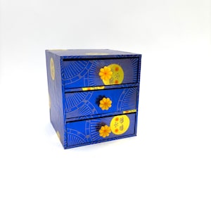 May include: A small, blue three-drawer chest with a celestial design. Each drawer features a yellow flower knob and a yellow moon and star design. The chest is decorated with a repeating pattern of lines and symbols.