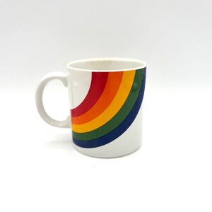Può includere: Una tazza in ceramica bianca caratterizzata da un motivo ad arcobaleno curvo e multicolore sul lato. Il design è composto da audaci bande a tinta unita di colore rosso, arancione, giallo, verde e blu che attraversano la superficie esterna.