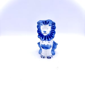 May include: A white ceramic lion figurine with blue and white painted details. The lion is sitting with its head held high and its mane is decorated with a blue and white pattern.