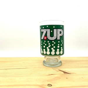 May include: A green glass 7 Up drinking glass with white polka dots and the 7 Up logo in white with white dots. The logo has the words "7 Up" in white with a small red "7 Up" logo in the upper left corner.