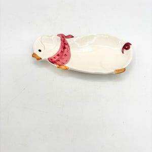 May include: A ceramic pig-shaped dish with a cream-colored body, a pink scarf, and brown accents. The dish has a small, shallow bowl for holding items. The pig's face is detailed with a small eye and a brown beak.