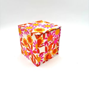 May include: A small square storage chest with three stacked drawers, featuring a vibrant floral pattern in shades of bright orange, hot pink, and yellow on a white background. Each drawer is equipped with a small, round, orange flower-shaped knob.