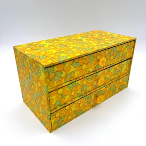 May include: A rectangular storage chest with three horizontal drawers, featuring a vibrant yellow and orange floral pattern with green leaves. Each drawer is equipped with a small, matching yellow flower-shaped knob for opening and closing.