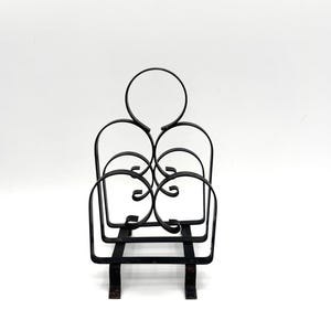 May include: Black wrought iron magazine rack with a decorative design. The rack features a series of curved and circular elements, creating an open, airy structure. The base has two feet for stability. The rack is designed to hold magazines or other reading materials.