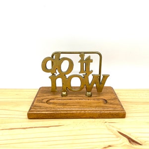 Op de afbeelding: Bureau-organizer van messing met een houten basis. Het messing deel heeft de tekst "do it now".