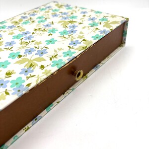May include: A rectangular box with a floral pattern in shades of blue, green, and white. The box has a brown interior and a small, gold-colored knob. The floral design covers the top and sides of the box.