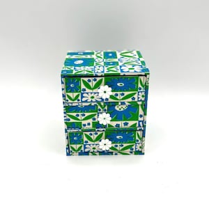 May include: A small three-drawer chest of drawers with a floral pattern in shades of blue, green, and white. Each drawer has a white flower knob.