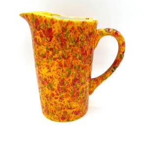 May include: A tall, ceramic pitcher with a yellow base and a speckled pattern of red, orange, and green. The pitcher has a curved handle and a spout for pouring. The interior is white.