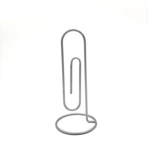 May include: A silver-colored metal desk accessory shaped like a large paperclip. The object features a vertical wire loop design that mimics the form of a standard paperclip, mounted securely onto a circular wire base for stability on flat surfaces.