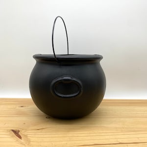 Puede incluir: Caldero de plástico negro con un asa de alambre. El caldero tiene un asa de anillo en el lateral.