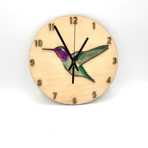 May include: A wooden clock with a hummingbird design. The clock face is light brown with gold numbers and black hands. The hummingbird is green, pink, and purple.