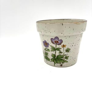 May include: A white ceramic flower pot with a speckled glaze and a floral design featuring purple, yellow, and green flowers.