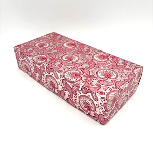 May include: A rectangular gift box with a silver and pink paisley pattern. The box is closed and has a shiny, reflective surface. The paisley design features a repeating floral motif in shades of pink and white.