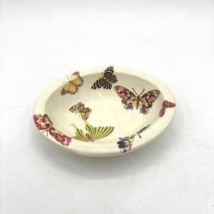 Może przedstawiać: Owalna, kremowa ceramiczna miska ozdobiona kolorowymi ilustracjami motyli. Motyle mają różne wzory i kolory, w tym żółty, fioletowy i czarny. Miska ma prosty, elegancki design, odpowiedni do serwowania lub ekspozycji.