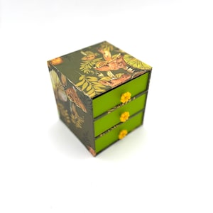 May include: A small, three-drawer box with a botanical design. The box has a dark green exterior with a pattern of mushrooms and foliage. The drawers are bright green with yellow flower-shaped knobs. The box is a cube shape.