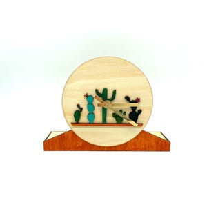 May include: A wooden clock with a cactus design. The clock face is round and has a light wood finish. The hands are gold colored. The clock is sitting on a wooden base.