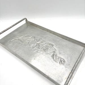 May include: A rectangular, silver-toned metal serving tray with a raised, embossed fruit and leaf design. The tray has a handle on one side and is likely used for serving food or drinks. The tray is made of metal.