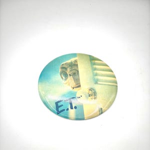 May include: Circular button with an image of E.T. peeking from a door. The button has a blue and yellow background with the text "E.T." in blue. A nostalgic collectible item.