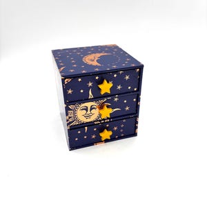 May include: A small dark blue storage chest with three drawers, featuring a celestial pattern of gold suns, moons, and stars. Each drawer is equipped with a bright yellow star-shaped knob, providing a contrast to the deep blue background.
