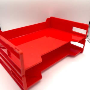 May include: A bright red plastic desk organizer featuring two stacked horizontal trays for document storage. The unit has an open-front design with side panels that include rectangular cutouts, providing a clear view of the tiered structure.