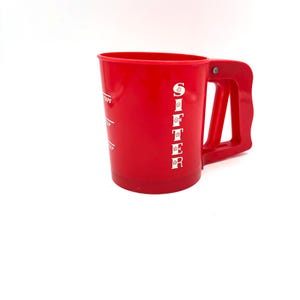 May include: A bright red plastic sifter with a handle. The word "SIFTER" is printed vertically on the side in white letters. The sifter also has measurement markings in white.