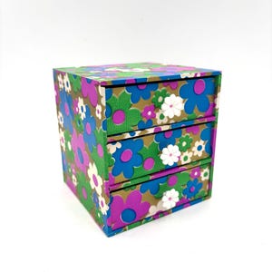 May include: A small, square, three-drawer storage chest covered in a vibrant floral pattern. The design features overlapping blue, purple, green, and white flowers on a gold background. Each of the three drawers has a matching floral front.