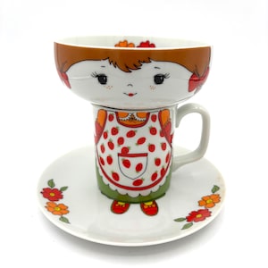 May include: A white ceramic cup and saucer set featuring a whimsical character design. The cup depicts a person with brown hair and a strawberry-patterned apron, while the saucer is decorated with small red and orange floral motifs.