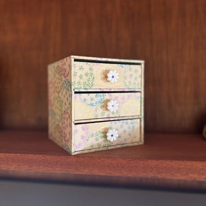May include: A small, three-drawer decorative box with a floral pattern in pastel colours. Each drawer has a white daisy-shaped knob. The box is made of cardboard and sits on a wooden shelf.