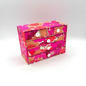 May include: A small, floral-patterned chest of drawers with four drawers. The chest is covered in a vibrant print featuring large pink, orange, and white flowers on a red background. The drawers have small, white flower-shaped knobs.