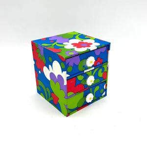 May include: A small, multi-coloured, floral patterned wooden box with three drawers. Each drawer has a white daisy-shaped knob.