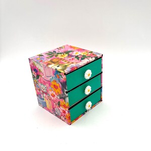 May include: A small, three-drawer dresser with a floral print featuring cats and flowers. The drawers are teal with white daisy-shaped knobs.