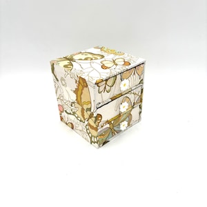 Peut inclure: Petite boîte carrée ornée d'un motif de papillons dans des tons beige, marron et jaune. La boîte comporte deux tiroirs visibles et un couvercle assorti. Le motif de papillon comprend des accents floraux.