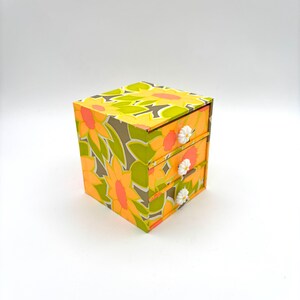 May include: A small, three-drawer box with a vibrant floral pattern in yellow, orange, and green. The box has white daisy-shaped drawer pulls. The design is reminiscent of vintage decor.